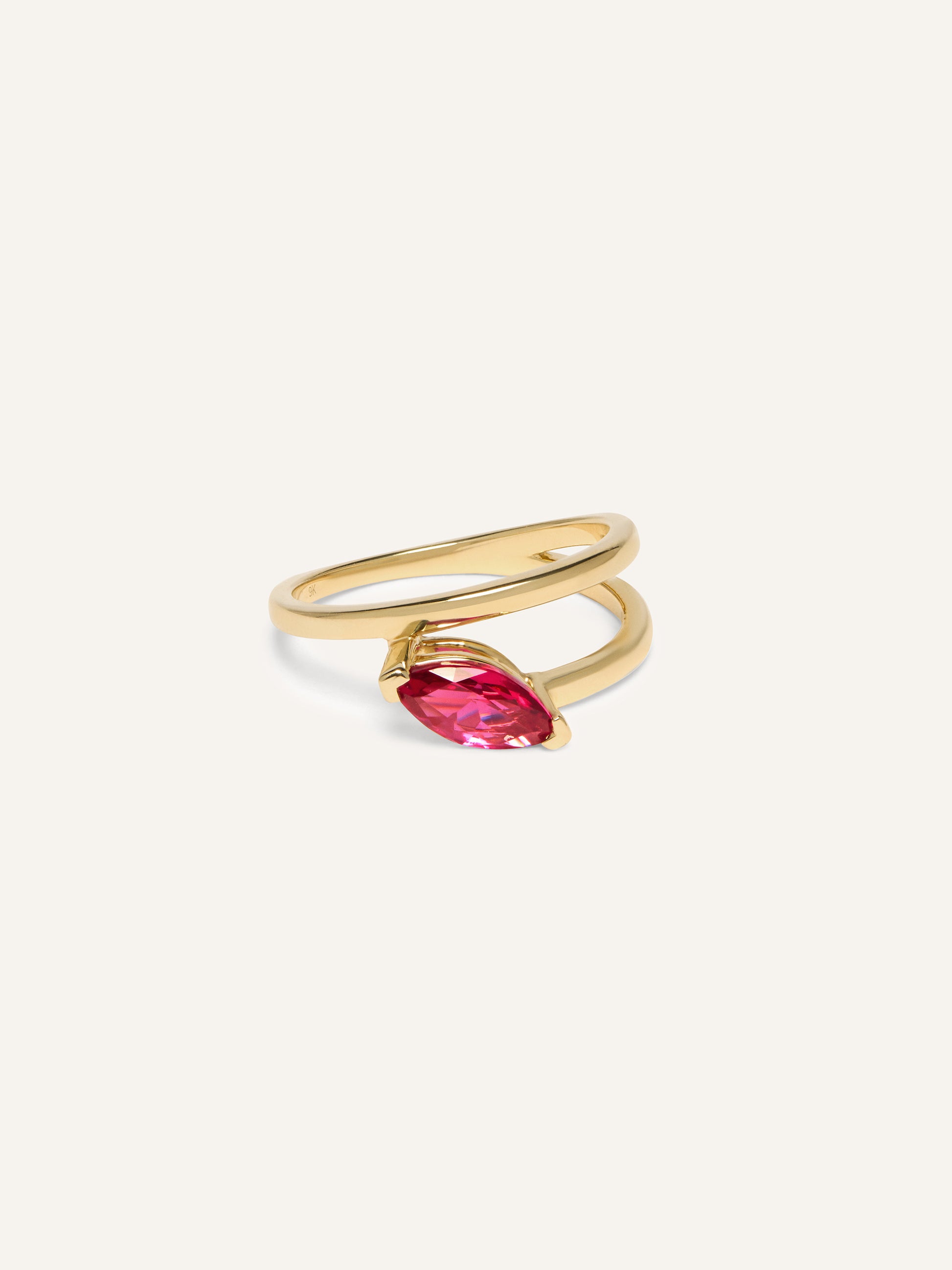 A gold double-band ring with a marquis ring with ruby gemstone, set diagonally, against a plain cream background. The tone is elegant and modern.